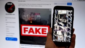 Në ekranin e telefonit shihet një ndërtesë e shkatërruar në Ukrainë, si pasojë e sulmit rus, ndërsa në monitorin e kompjuterit, një faqe online që përhap lajme të rreme.